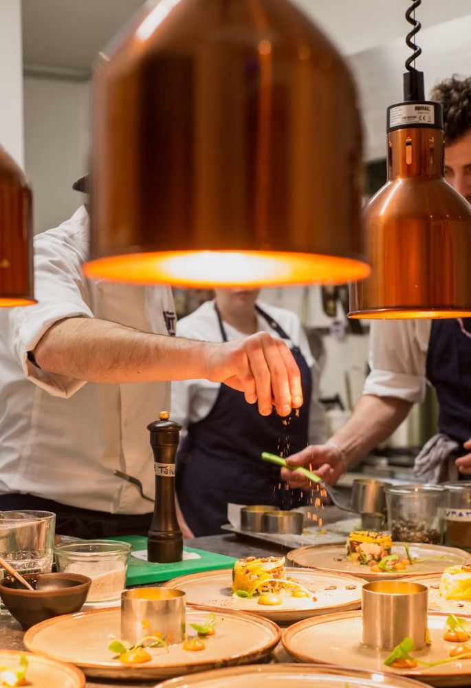 A chef finishing plates under copper heat lamps in a working kitchen